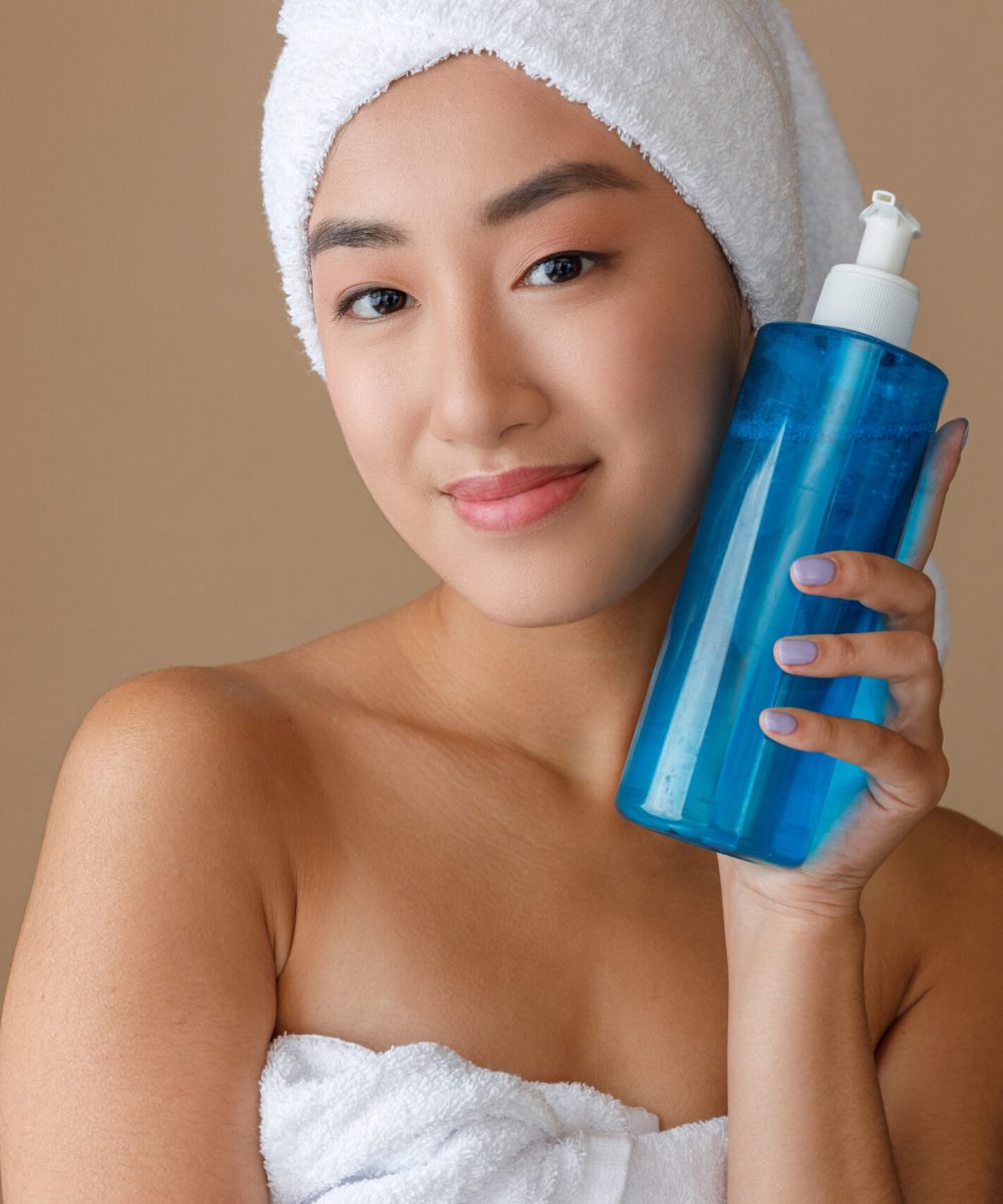 Smiling female person with moisturizing lotion in her hand standing in studio. Isolated on light brown background